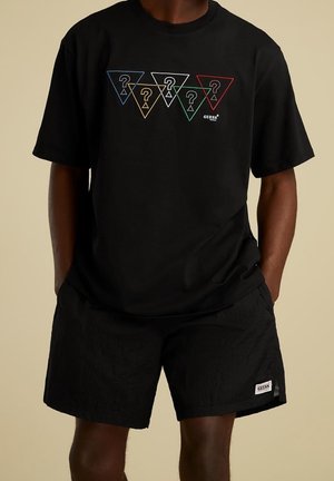 Homme portant un t-shirt noir avec cinq logos en forme de point d'interrogation triangulaires colorés au-dessus du texte blanc "GUESS", associé à un short noir avec étiquette.