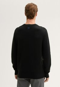 Schwarzer, gestrickter Pullover mit rundem Halsausschnitt, langen Ärmeln und einem kleinen Logotag am Nacken. Das Material wirkt weich und strukturiert.