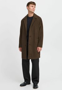Brun ullblandings overshirt med slippkrage, frontknapper og to sidelommer. Brukt over en svart t-skjorte og svarte bukser.