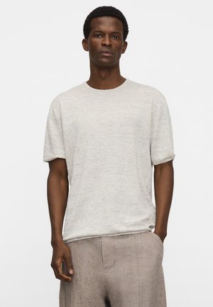Mann trägt hellgraues kurzärmliges T-Shirt und hellbraune Hose, steht mit einer Hand in der Tasche vor weißem Hintergrund.