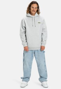 Sudadera gris con capucha, que cuenta con un bolsillo frontal y un pequeño logo verde. Combinada con unos vaqueros holgados de color azul claro y zapatillas blancas.
