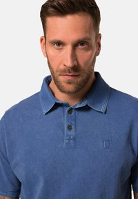 Modrá polo tričko z jemné tkaniny, s klasickým límečkem, zapínáním na tři knoflíky a vyšitým logem na levé hrudi.