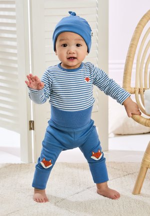 Malé dieťa v modrom klobúku, pruhovanom tričku a nohaviciach s líščími záplatami, stojí bosé a drží prútenú stoličku v svetlej miestnosti.