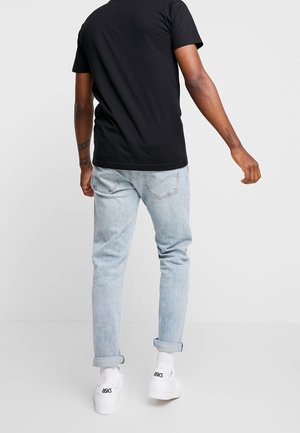 Homme portant un t-shirt noir, un jean bleu clair retroussé, des baskets blanches Asics, debout de dos sur un fond gris uni.