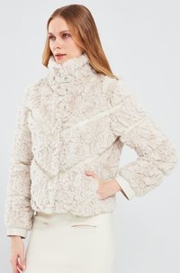 Nep bveroos in lichtbeige met een hoge kraag, textuur oppervlak en contrasterende gladde afwerking. Voorzien van een ritsluiting en brede mouwen.