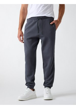 Uomo che indossa pantaloni jogger grigio scuro con vita con coulisse e sneakers bianche, in piedi su sfondo bianco.