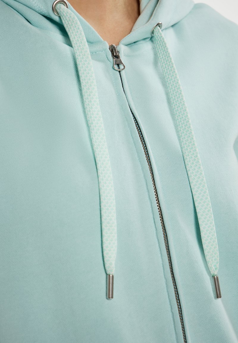 Felpa cappotto azzurro chiaro con zip, dotata di lacci decorati, accessori in metallo argentato e una texture morbida in misto cotone.