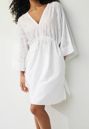 Robe blanche avec un col en V, des manches courtes et un motif en dentelle ajourée. Le tissu est léger avec une taille froncée et des fentes latérales.