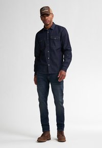 Camisa de mezclilla oscura con dos bolsillos en el pecho, combinada con jeans azules y botas marrones. Una gorra beige con un logo completa el atuendo.