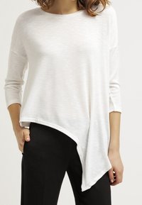 Asymmetrisches weißes Oberteil mit strukturiertem Finish, lange Ärmel und asymmetrischer Saum. Kombiniert mit schwarzen Hosen.