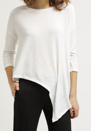 Top bianco asimmetrico con finitura testurizzata, caratterizzato da maniche lunghe e un orlo drappeggiato. Abbinato a pantaloni neri.