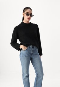 Juodas megztinis su aukštu kaklu ir trumpomis rankovėmis, derinamas su šviesiai mėlynomis aukštu liemeniu džinsais. Modelis dėvi saulės akinius.