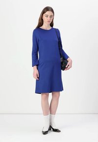 Sininen, pitkähihainen mekko, pyöreällä pääntiellä, rento istuvuus, ylettyen puoliväliin reittä. Yhdistetty valkoisten sukkien ja mustien korkokenkien kanssa.