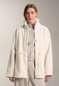 Giacca in pile color crema, soffice, con collo alto, chiusura frontale a clip e due tasche laterali con zip. Interno liscio.