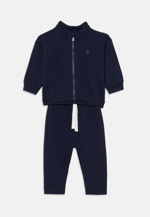Mørkeblå joggedress med glidelåsjakke og lange bukser. Mykt stoff, høy krage og hvit snor på buksene. Enkelt, ensfarget design.