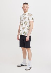 Kortermet poloskjorte med palme-mønster i beige og grønt, kombinert med svarte shorts og hvite joggesko med svarte striper.