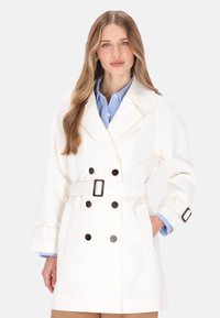 Hvid trenchcoat med dobbeltradet front, store revers, bælte i taljen og brune knapper. Har opkrogede ærmer og sidelommer.