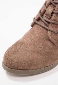 Bota de ante marrón para tobillo con punta redondeada, detalles de cordones y suela de goma texturizada. El material parece suave y mate.