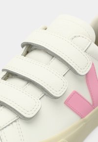 Άσπρο αθλητικό παπούτσι με τρεις λουρίδες βέλκρο και ροζ λογότυπο "V" στο πλάι, με μπεζ σόλα και ραμμένες λεπτομέρειες.