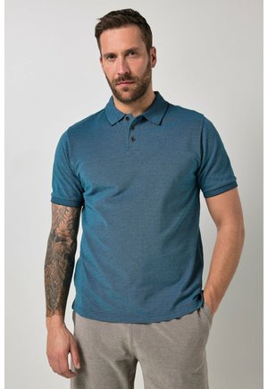 Homme aux cheveux courts et foncés avec une barbe, portant un polo manches courtes bleu sarcelle et un pantalon gris, debout avec une main dans la poche.