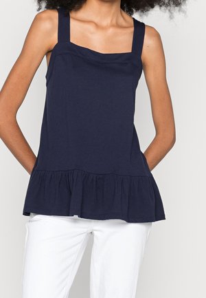 Kobieta w granatowym topie bez rękawów z szerokimi ramiączkami i falbaną u dołu, zestawionym z białymi spodniami na jednolitym tle.