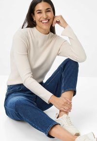 Maglione a collo alto rigato color crema, jeans blu rilassati e sneaker chiare. Posizione seduta con la mano appoggiata sul ginocchio, sfondo neutro.
