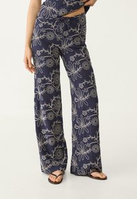 Navy losse-fitting broek met witte bloemenborduring, voorzien van een trekkoord in de taille, gedragen met zwarte teenslippers en een cropped top, met zichtbare benen en hand.