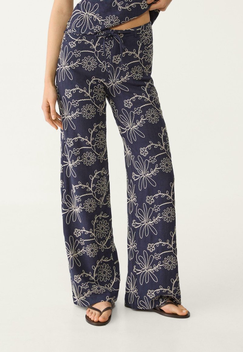 Navy losse-fitting broek met witte bloemenborduring, voorzien van een trekkoord in de taille, gedragen met zwarte teenslippers en een cropped top, met zichtbare benen en hand.