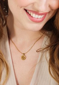 Gouden ketting met een ronde hanger met een gestileerde "F" in een contrasterende kleur, rustend op een textuurstof van lichte kleur.