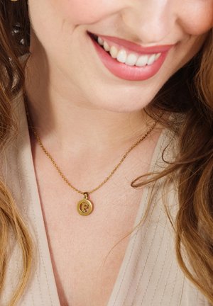 Gouden ketting met een ronde hanger met een gestileerde "F" in een contrasterende kleur, rustend op een textuurstof van lichte kleur.