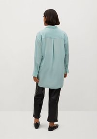 Chemise bleu clair surdimensionnée avec col, manches longues et un empiècement au dos, associée à un pantalon en cuir noir et des chaussures foncées.