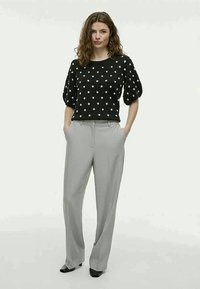 Woman standing with hands in pockets wearing black polka-dot sweater, light gray wide-leg trousers, and black shoes.