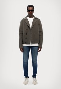 Cappotto peacoat grigio in lana con ampi revers e bottoni neri; indossato sopra una camicia bianca. Jeans skinny blu e sneakers bianche completano il look.