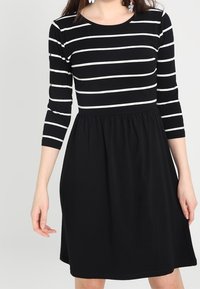 Robe noire à manches trois-quarts, avec des rayures blanches horizontales sur la moitié supérieure et une jupe froncée. Tissu lisse avec une coupe décontractée.