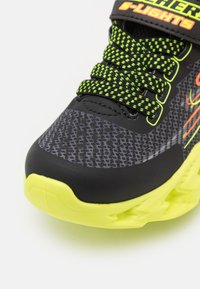Black and gray athletic shoe with a textured upper, neon green laces, and a bright yellow sole. Features light accents and a secure strap.