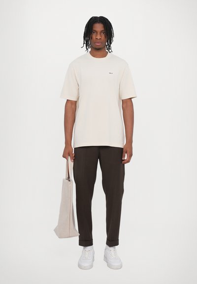 Camiseta de algodón beige con mangas cortas y pequeño logo, combinada con pantalones ajustados marrón oscuro y zapatillas blancas, sosteniendo una bolsa de tela clara.