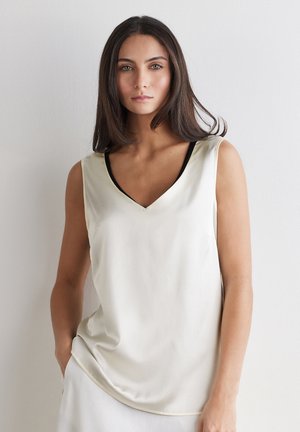 Junge Frau mit glatten dunklen Haaren, die ein ärmelloses cremefarbenes Top mit V-Ausschnitt und schwarzer Unterlage trägt, steht vor einer weißen Wand.