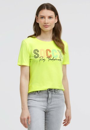 Young woman with long brown hair wearing a bright yellow T-shirt with "SOCCX Reg. Trademark" text and light gray jeans against a plain background.