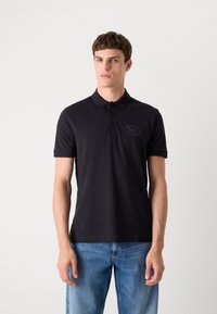 Camisa polo preta com gola texturizada, três botões e um pequeno logótipo tonal no peito. Usada com jeans de ganga azul-claro.