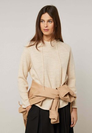 Frau mit langen braunen Haaren, die einen beigen, gerippten Hoodie und einen schwarzen Rock trägt, mit einer beige Jacke, die um ihre Taille gebunden ist, vor neutralem Hintergrund.