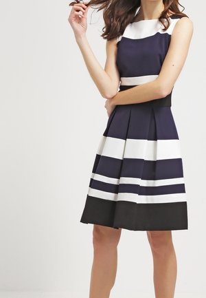 Vrouw die een mouwloze donkerblauwe jurk draagt met brede, witte en zwarte horizontale strepen, staand met één arm gekruist en de andere hand die het haar aanraakt.