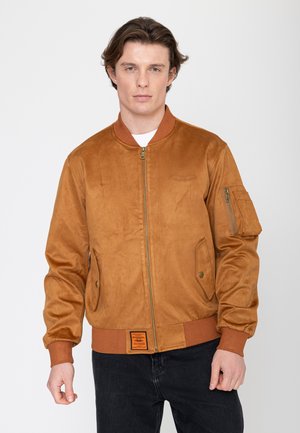 Jeune homme à la peau claire et aux cheveux bruns portant une veste aviateur en daim marron et un pantalon noir, debout devant un fond blanc.