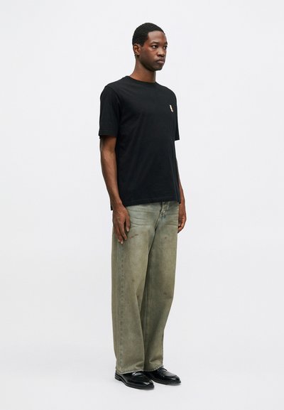 Hombre de pie vistiendo una camiseta negra con un pequeño logo en el pecho, jeans de lavado claro anchos y zapatos negros, contra un fondo blanco plano.