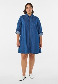 Kurvig kvinna som bär en jeansskjortklänning med uppvikta ärmar och vita skor, stående mot en enkel vit bakgrund.