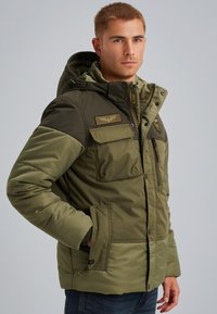 Grön vadderad jacka med huva, utrustad med två framfickor, dragkedja och en patch på bröstet. Strukturerat tyg och kontrasterande ärmar.