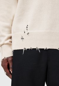 Beiger Strickpullover mit geribbeltem Saum und ausgefransten Details. Über schwarzem Stoff drapiert. Sichtbares Ausfransen und kleine Risse am unteren Rand.