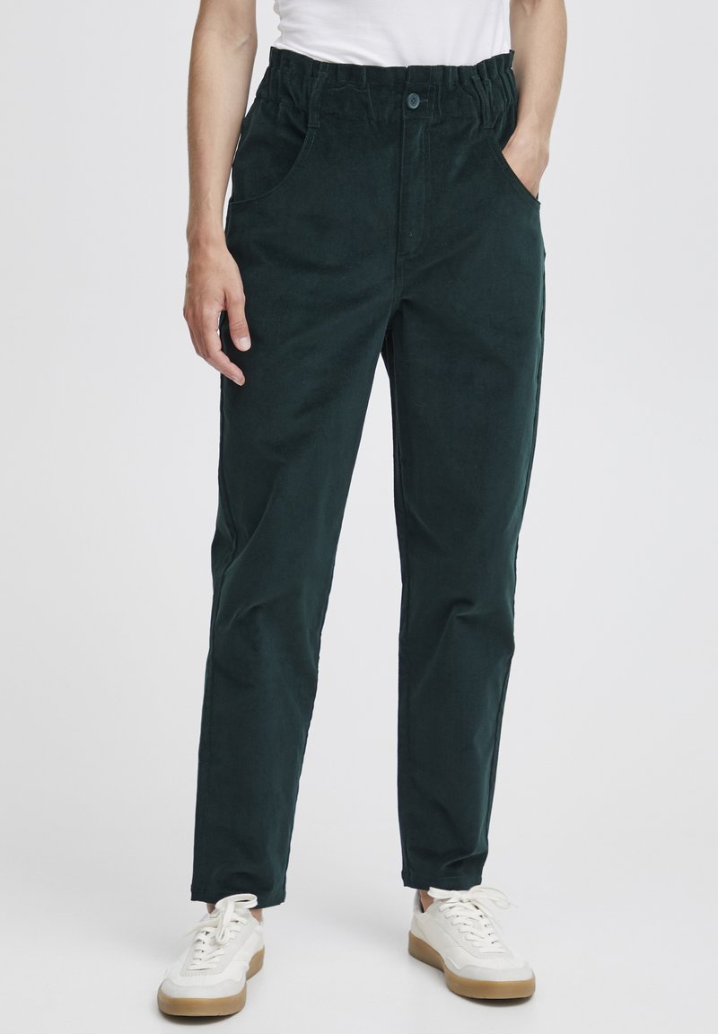 Dark green corduroy trousers with an elastic waistband, side pockets, and a straight-leg cut, paired with white sneakers.