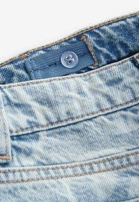 Jean en denim bleu clair avec une finition texturée, présentant des coutures, un patch en tissu bleu et un bouton en plastique transparent à la taille.