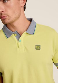 Polo shirt jaune clair avec un col texturé gris, arborant un petit logo en patch sur la poitrine gauche et des boutons d'accent bleu.