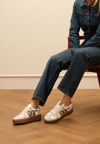 Weißes Sneakers mit brauner Sohle, versehen mit einem Leopardenmuster-Akzent. Kombiniert mit blauen Jeans in einer Sitzposition.